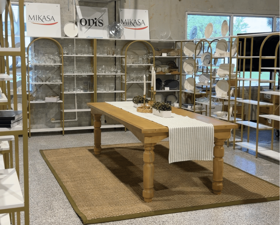 wood farm table surrounded by shelves with glassware and plates.  Mikasa and Onis signs 