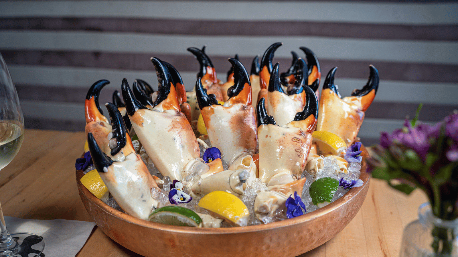A brown bowl of Florida stone crabs standing up with claws on top, Inside the blow is slices of lemon, lime and some flower petals.