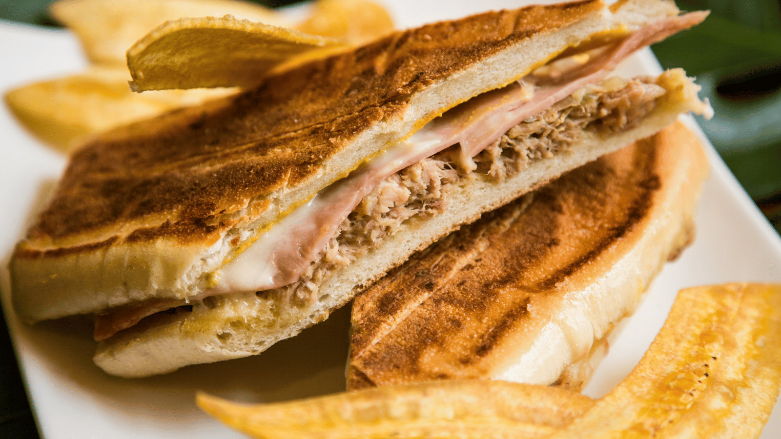 Pressed Cuban sandwich sliced in half, wih fried plantains surrounding it.