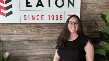 Woman in black shirt and with glasses stands in front of wood wall with Eaton Marketing logo on it. 