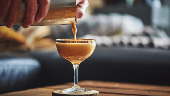 Martini glass on wood pedestal being filled with espresso martini with shaker above