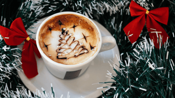 Small cup filled with espresso and topped with foam that has a Christmas tree decorated on top. Cup sits on a white saucer that is surrounded with green and white garland that has two red bows on it.