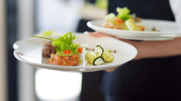 Two white plates are being held by one hand and wrist. The plate in the front, held by the hand, has two food items placed on it that look like crab cakes. Next to the crab cakes are thin slices of cucumber filled with a creamy cheese mixture. The plate in the back is resting on a wrist and topped with food items that look similar to the first plate.