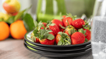 Three green plates with brown trim stacked on top of each other. Strawberries are on top with a pitcher of green smoothie and an orange in the background.