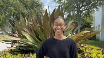 Woman with brown skin and eye glasses, wearing a black long sleeved shirt and smiling standing in front of large plant