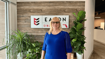 Woman in blue blouse and glasses stands in front of wood wall with Eaton Marketing logo