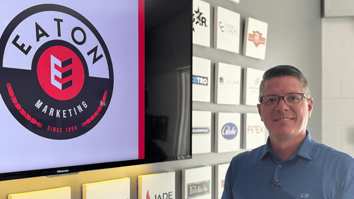 Man with salt and peper hair wearing glasses and a blue collared shirt stands to the left of a large Eaton Marketing logo on a wall that's surrounded by smaller logos of different foodservice equipment brands.