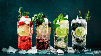 Four glasses filled with ice and different color cocktails. A slice of lime are in three of them and all glasses are garnished with mint and rosemary. 