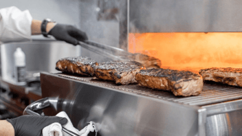 Chef with white coat and black gloves holding tray while other hand flips steaks under electric broiler