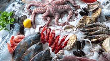 A variety of different types of seafood, including octopus, crawfish, shrimp, whole fish, and mussels, on a display of shaved, or flake ice. A lemon and sprig of parsley are also shown.