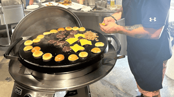 Evo wheeled grill with hamburger patties and buns cooking on top. To the left chef's arms and hand in view, holding cheese to put on patties. 