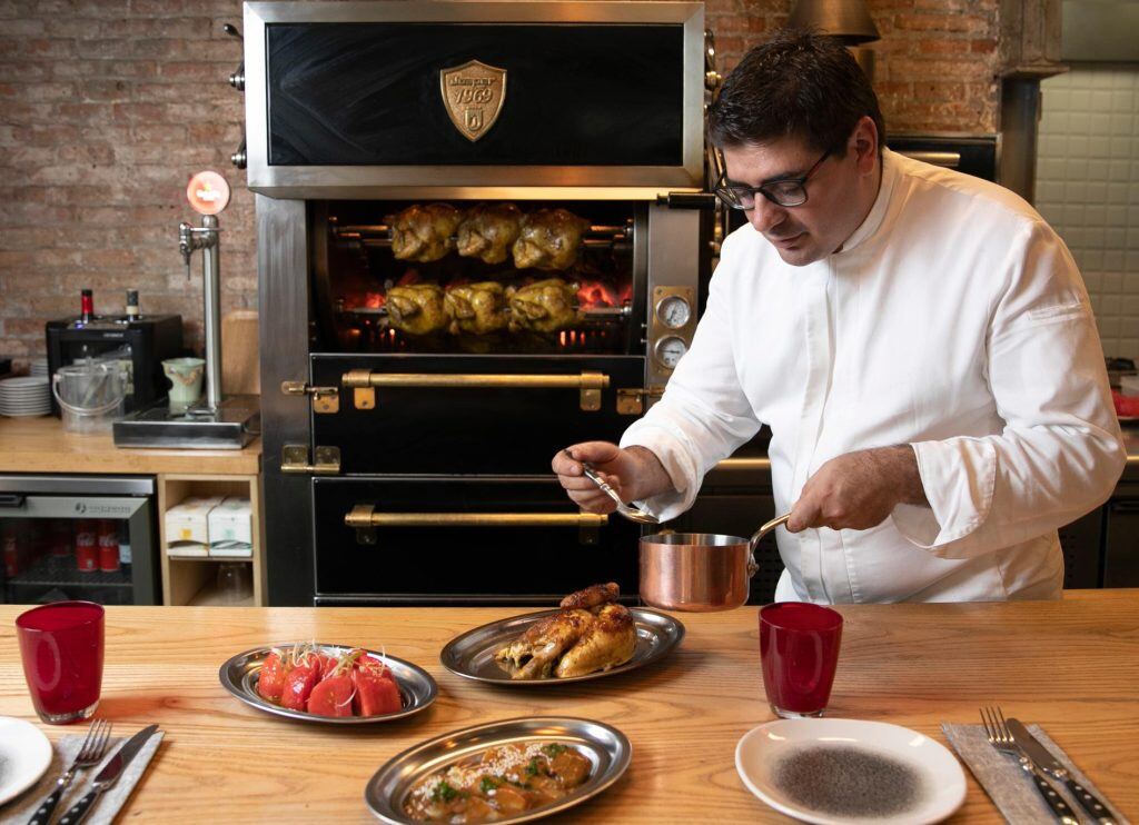 Chef plating meals on silver chargers and spooning sauce over one. In background is Josper Rotisserie with six chickens inside.