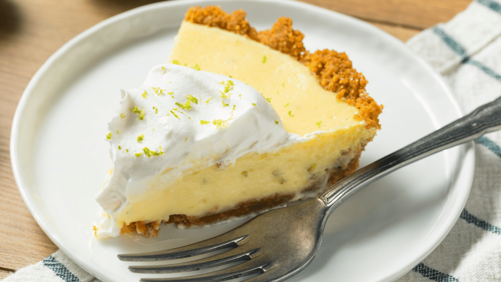 Slice of key lime pie on a white plate. A fork sits next to the pie, which has a dollop of whipped cream with lime zest on the tip of the pie.