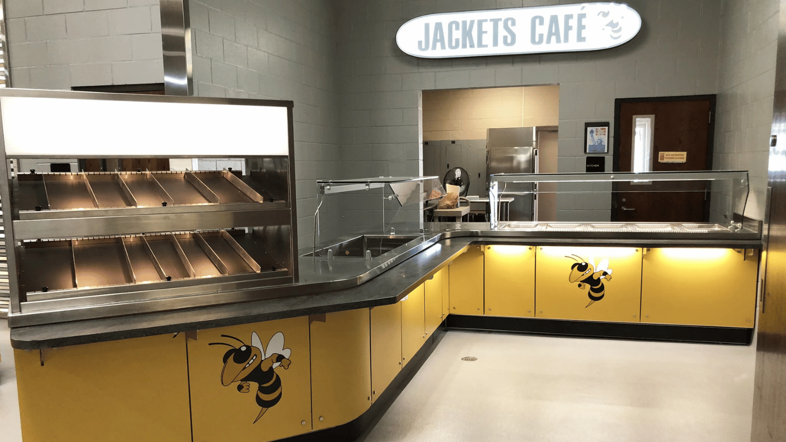 View of a high school serving counter. Bottom part of counters are yellow with two panels having yellowjackets on them. Above on wall is sign that says Jackets Cafe.