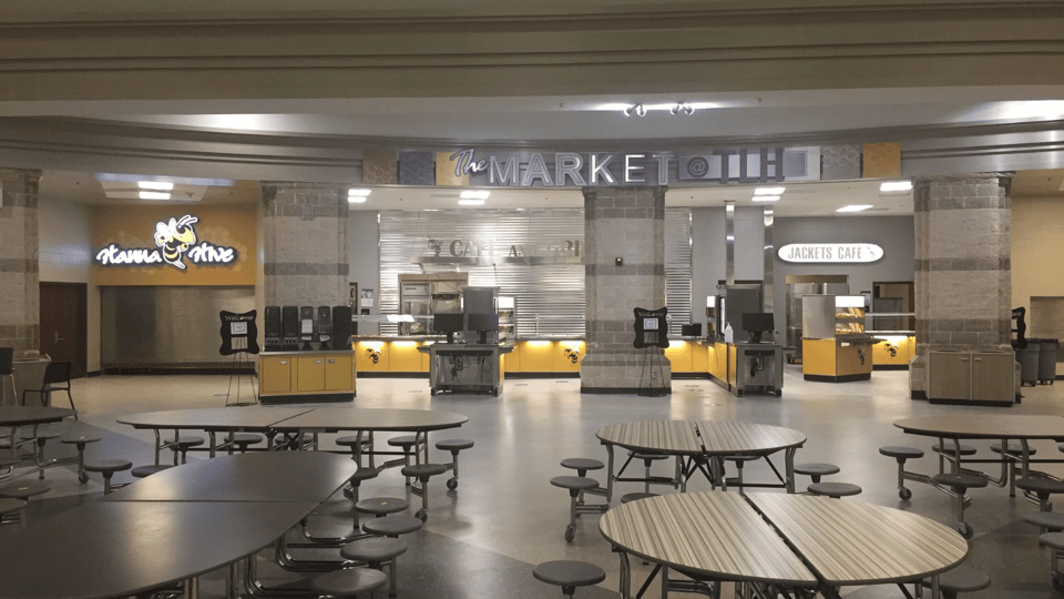 Wide view of high school cafeteria. In foreground are round tables with attached stools. In background is LTI counters and point of sale stations. Metal sign above says Market and lit sign on wall to right has large black and yellow yellowjacket mascot.