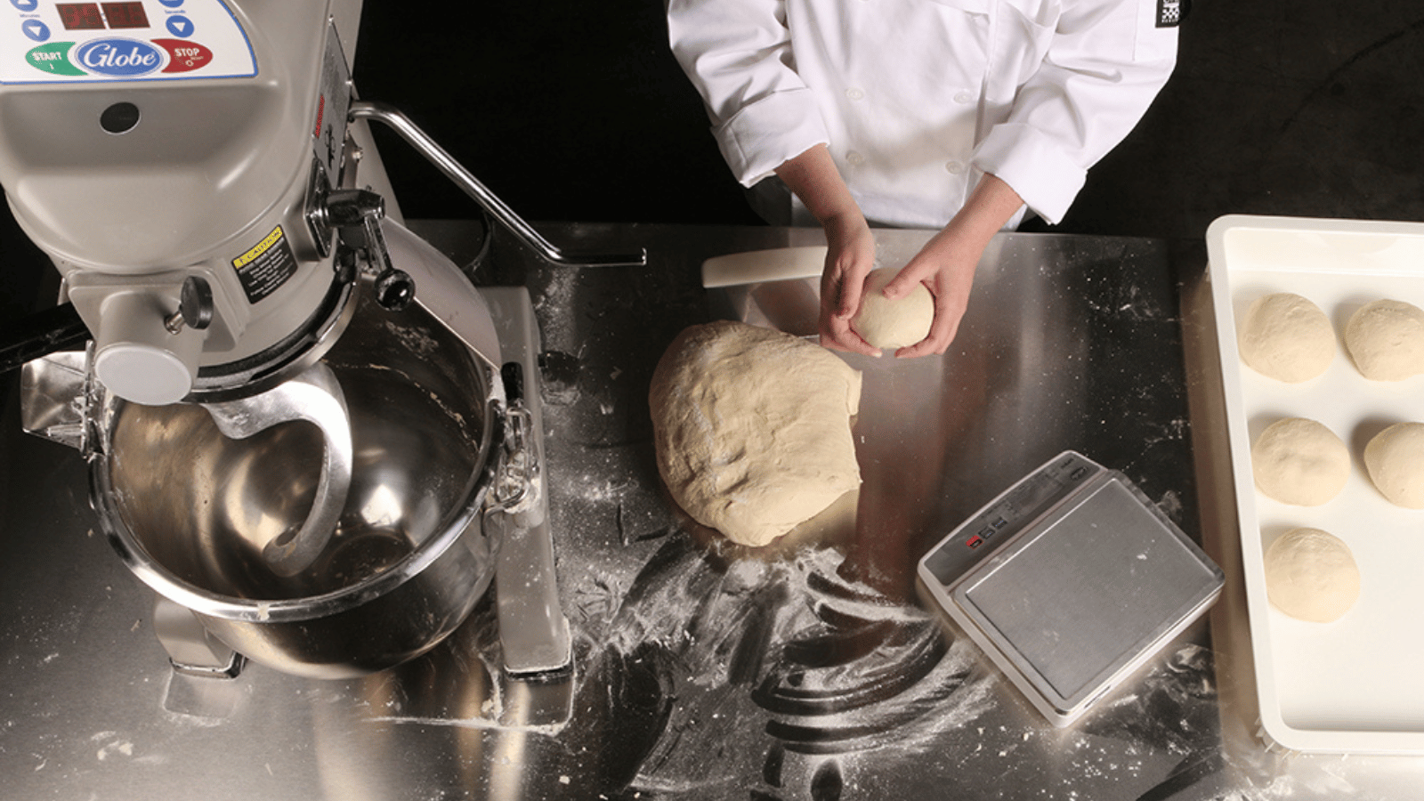 Overview of a Globe commercial mixer. Next to is is the torso and arms of a person in a white chef's coat with a raw dough ball in their hands. In front of them on a stainless steel table with flour is a larger ball of raw dough and a food scale. Next to the food scale are portioned out dough.