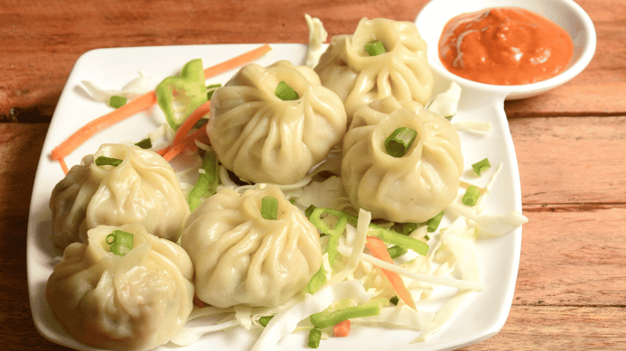 Six steamed soup dumplings on a white plate surrounded by shavings of carrots and cabbage. Small bowl of red dipping sauce sits to the top right of plate.