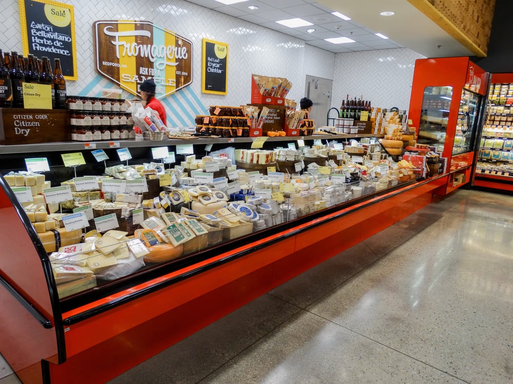 View of long red refrigerated display case in a grocery store. Case is full of assortment of different cheese. Wall has a sign that says 'Fromagerie.'