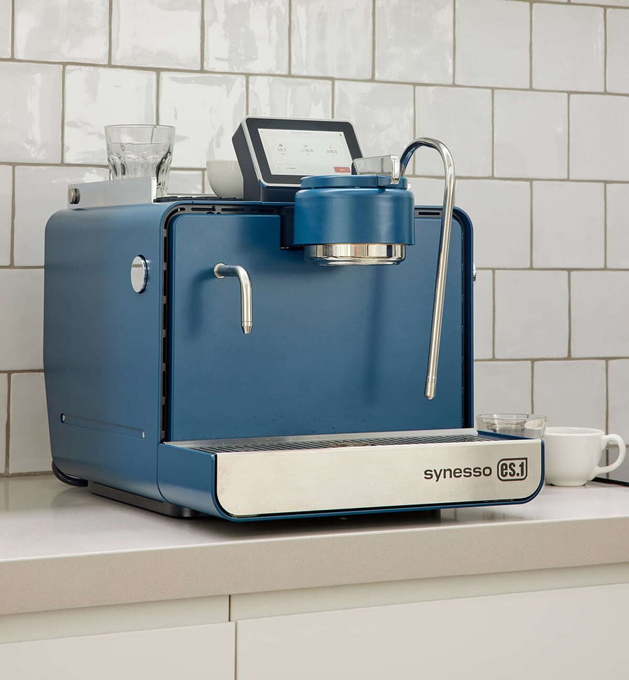 Steel blue Syneso ES.1 espresso machine sits on steel countertop with white cup next to it. Behind machine is white tile wall. 