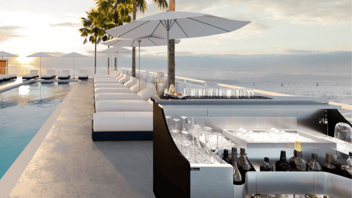 A view of an outdoor patio at a hotel. There's a stainless-steel mobile bar on the right side of long inground pool filled with sparking blue water. Behind the bar, to the right of the pool is a long row of white luxury lounge chairs. There are tall white umbrellas above the white lounge chairs.