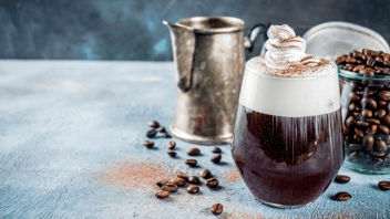 A stemless glass of a hot espresso martini topped with whipped cream slowly melting into the coffee and over the rim of the glass. Behind the glass is a silver pitcher. There are 20 coffee beans scattered on a table in front of the pitcher and glass which are also placed on a gray table. 