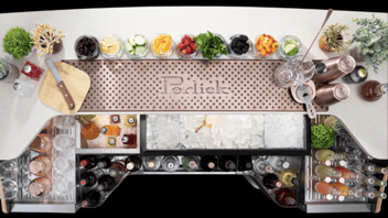 An overhead view of a mobile bar unit. From above, the top of a silver bar  with the word Perlick pressed into a long perforated drain across the top of the bar. There are ten small glass cups lining the outside of the drain and each cup contains a different kinds of fruits and condiments for alcoholic drinks.