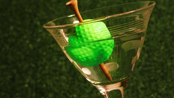 Martini glass half full with clear liquid and ice cube holder shaped like green golf ball with tee through the top.