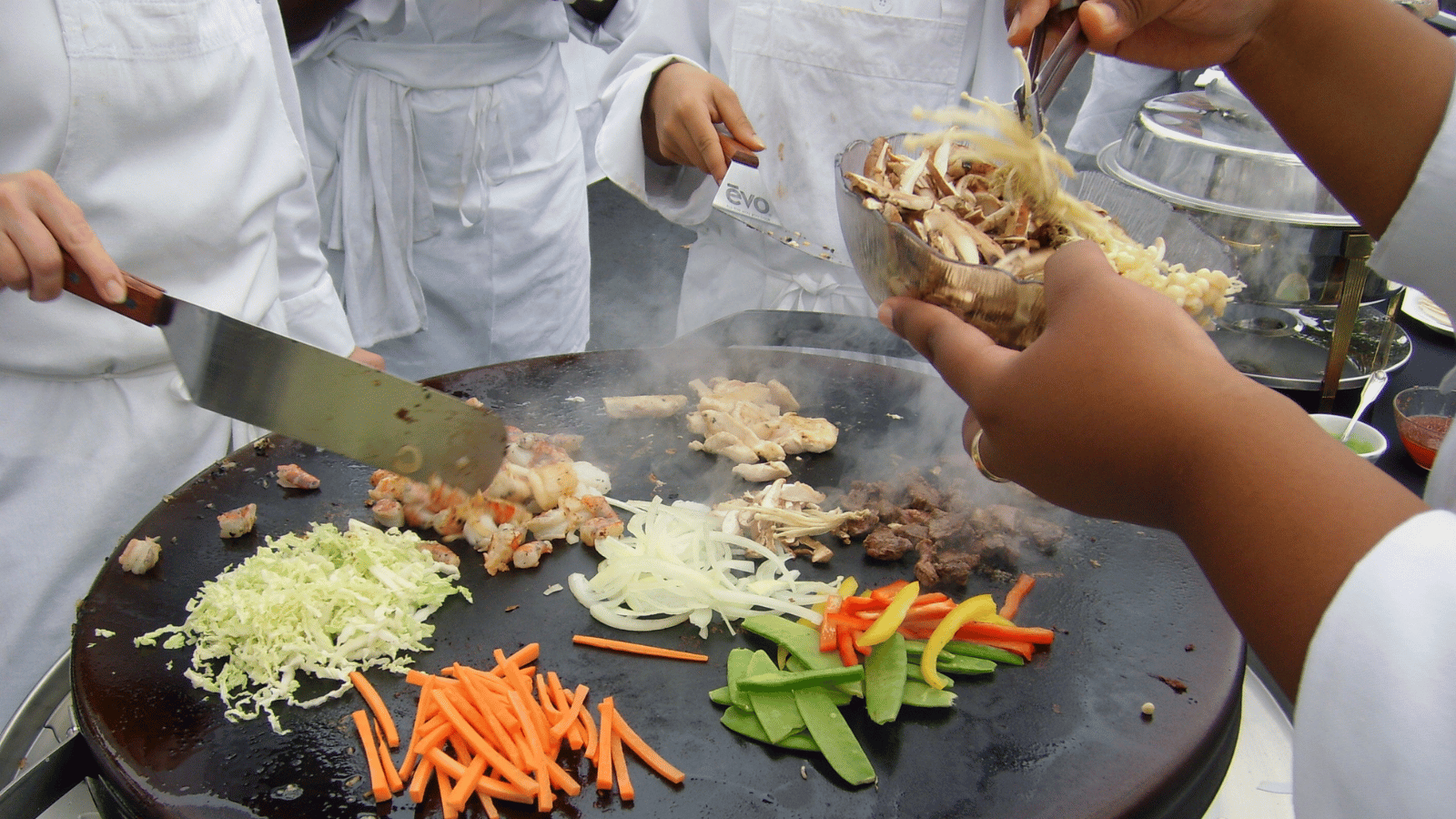 Close up of Evo EVent ventless griddle with matchstick carrots, sliced onions, pea pods, and steak being cooked on top