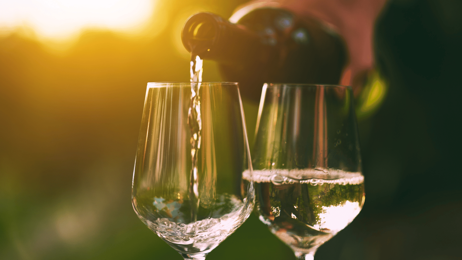 Two glasses in close up. One has half glass of white wine while wine bottle pours white wine in the other glass. Sunset is in the background.