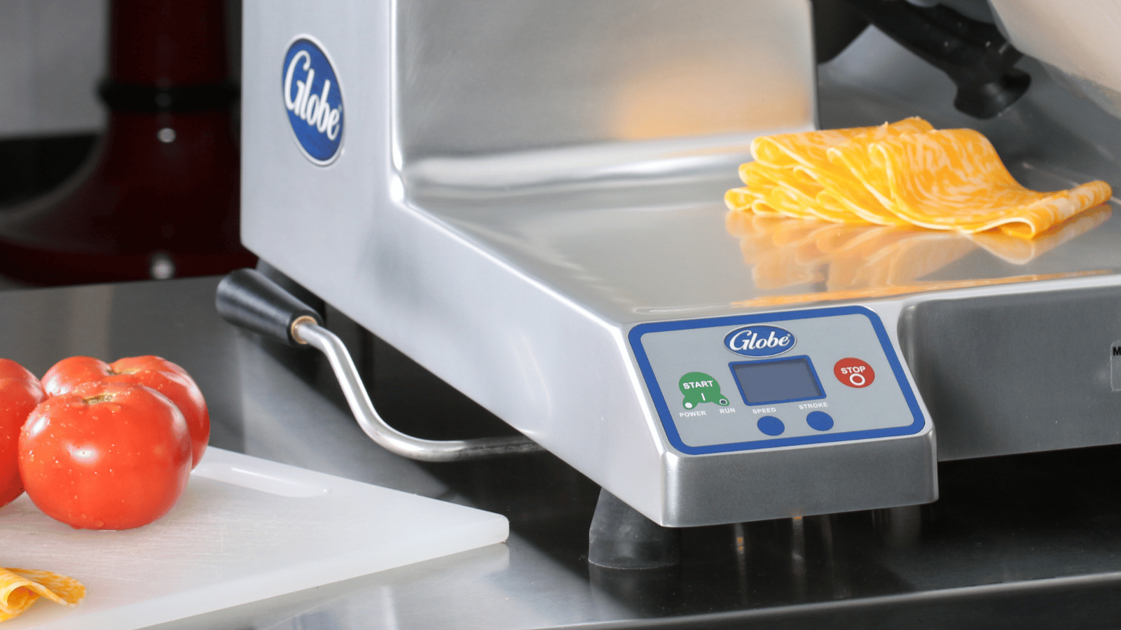 Upclose image of Globe slicer with sliced Colby Jack cheese on it. Next to the slicer are whole tomatoes on a plastic cutting board. 