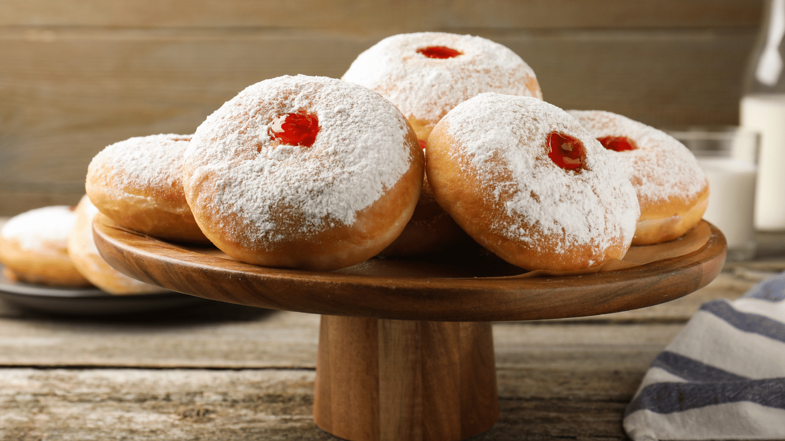 Five fried donuts with dollops of jelly, covered in powdered sugar sit on wood pedestal. Blue striped towel to side and glass of milk in background.