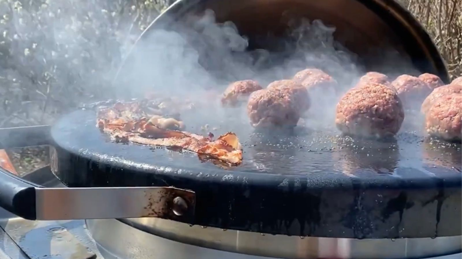 Close up photo of a mobile flattop commercial grill. The top of the grill is round and black. There are multiple strips of bacon, and a bunch a very large meatballs cooking on top of the grill. Steam is rising up from the sizzling meatballs and bacon.